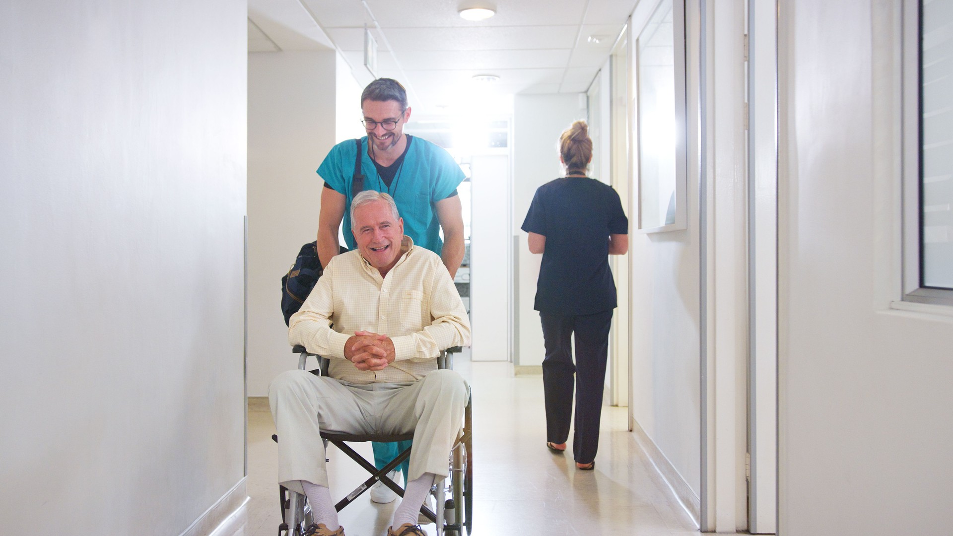 Male Orderly Pushing Senior Male Patient Being Discharged From Hospital In Wheelchair